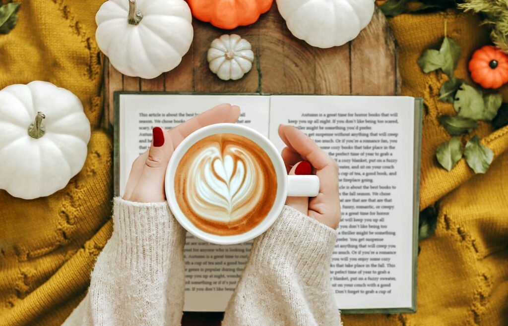 A woman's hands holding a coffee with a book on a table decorated for fall with ceramic pumpkins.