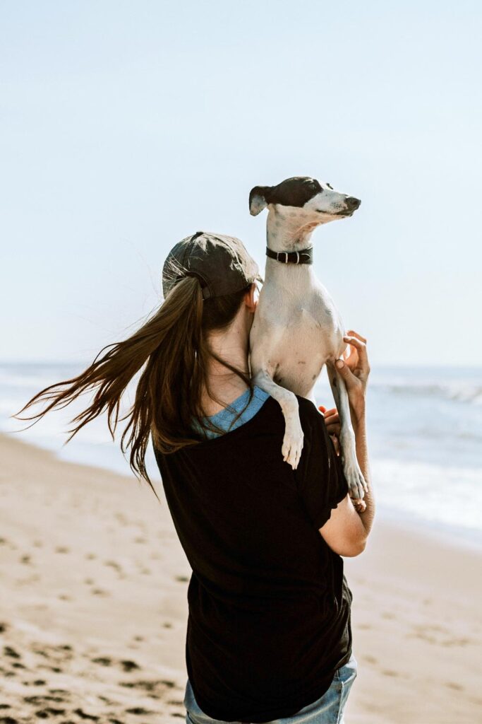 A woman holding a dog on her shoulder at the beach.