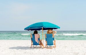 Two women sitting in chairs on the beach under an umbrella.