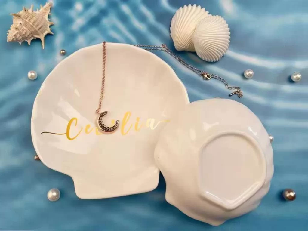 A white ring dish shaped like a scallop shell, with someone's name in gold script..