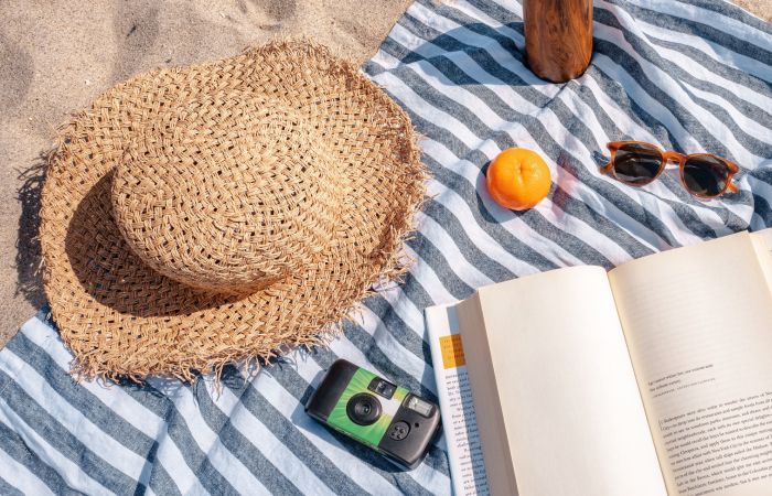 A beach blanket with a sun hat, book, drink, sunglasses, and an orange.