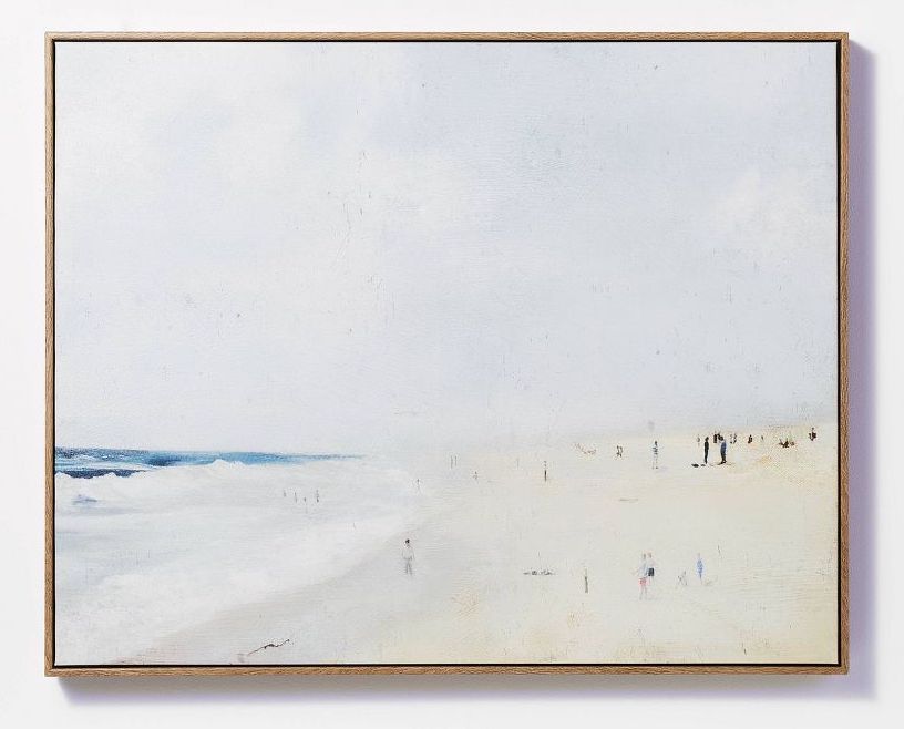 A framed canvas painting of people on the beach and the ocean waves. The sky is hazy.