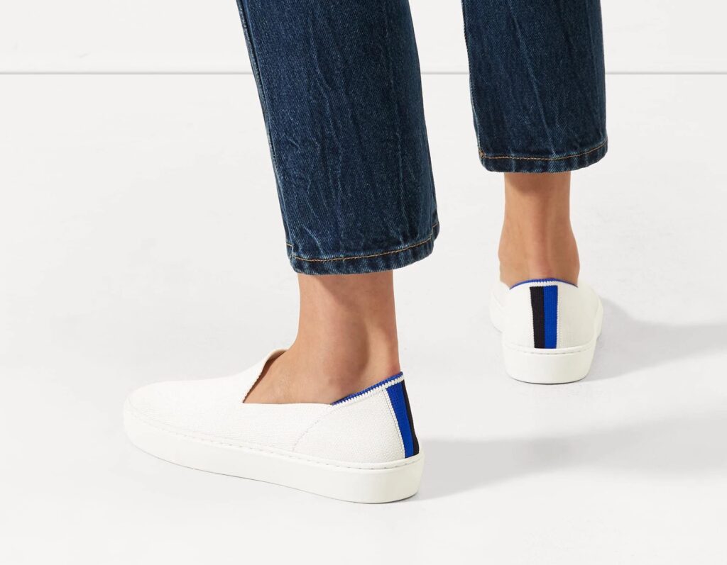 The legs and feet of someone wearing a pair of jeans and a pair of white slip-on sneakers with a blue and navy stripe on the heel.