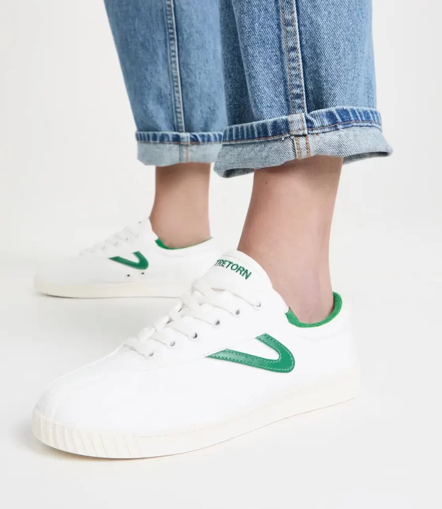 The legs and feet of a person wearing a pair of jeans and a white and green pair of Tretorn sneakers.