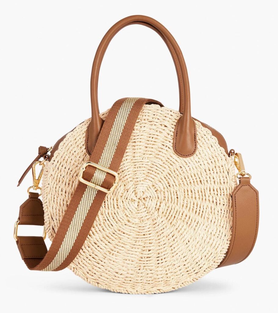 A round woven straw tote bag with brown handles and a crossbody strap.