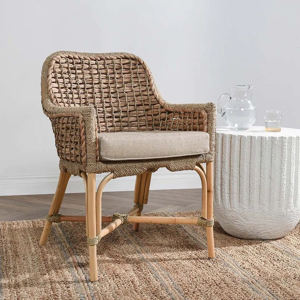 A rattan chair in earth tones with a cushion, shown in a room next to a side table.