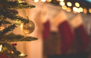 A gold ornament hanging from the branch of a Christmas tree. In the background, out of focus, stockings are hanging from a fireplace mantel.