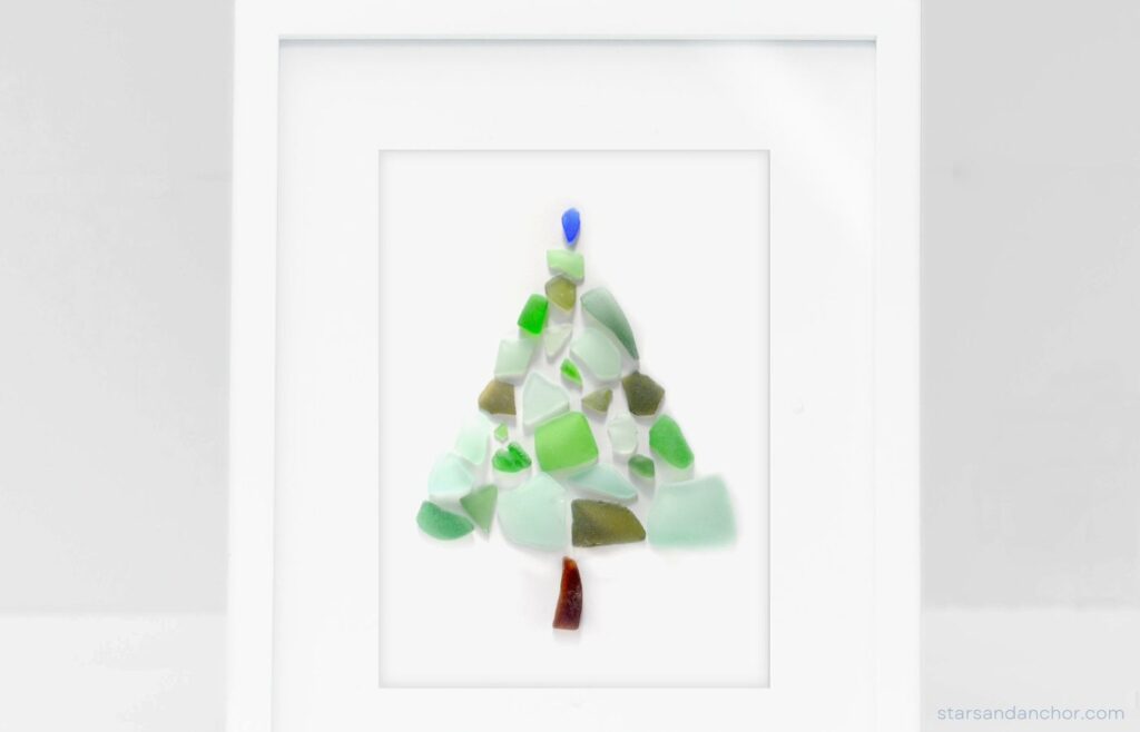A sea glass Christmas tree made out of green, blue, and brown sea glass in a white picture frame.