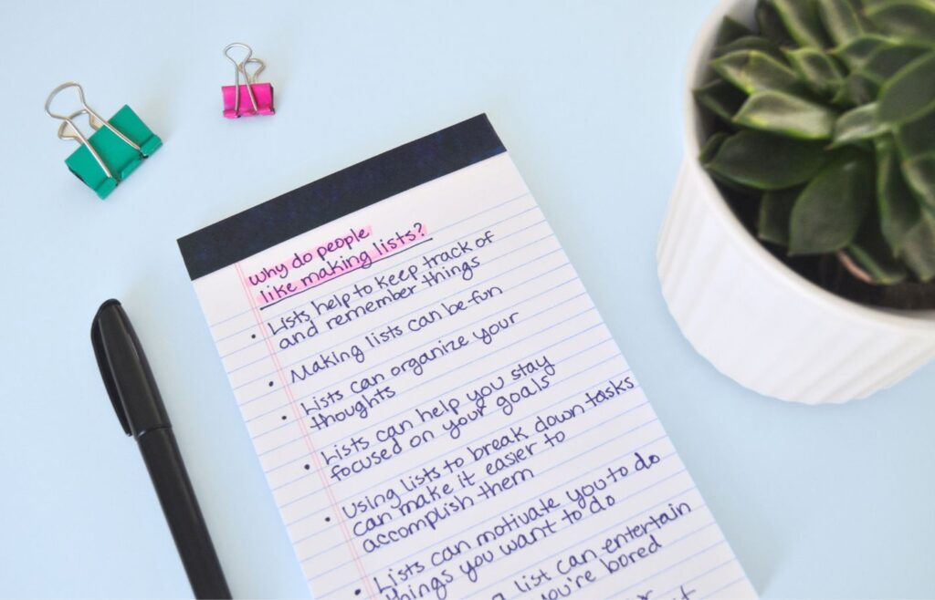 Binder clips, a pen, a plant, and a notepad on a desk. The notepad says, why do people like making lists? With some items bulleted underneath, that are listed below in this article.