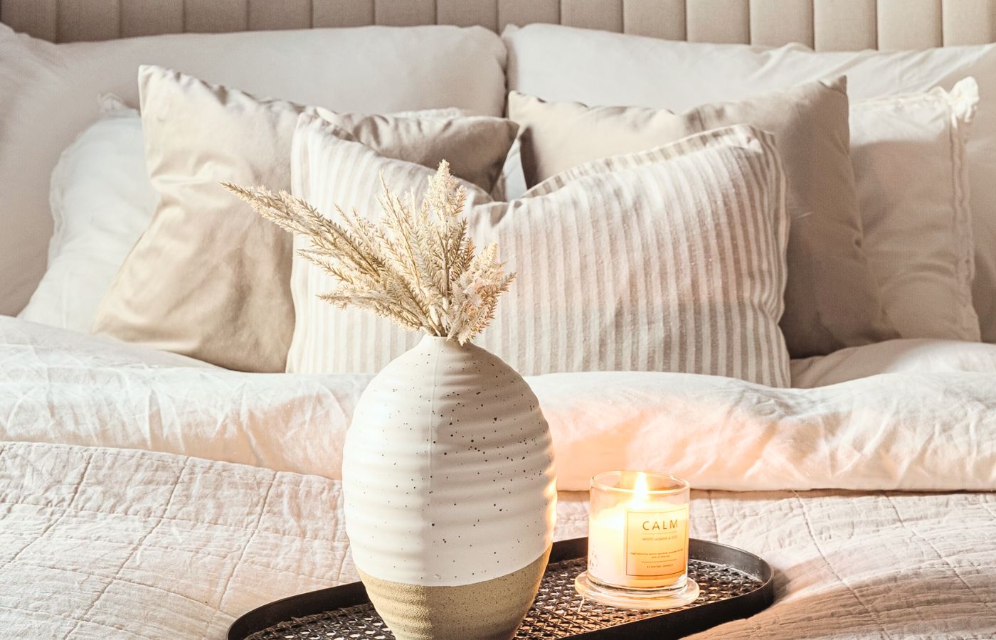 A bed with beige linens and a tray containing a vase with dried grasses, and a candle.