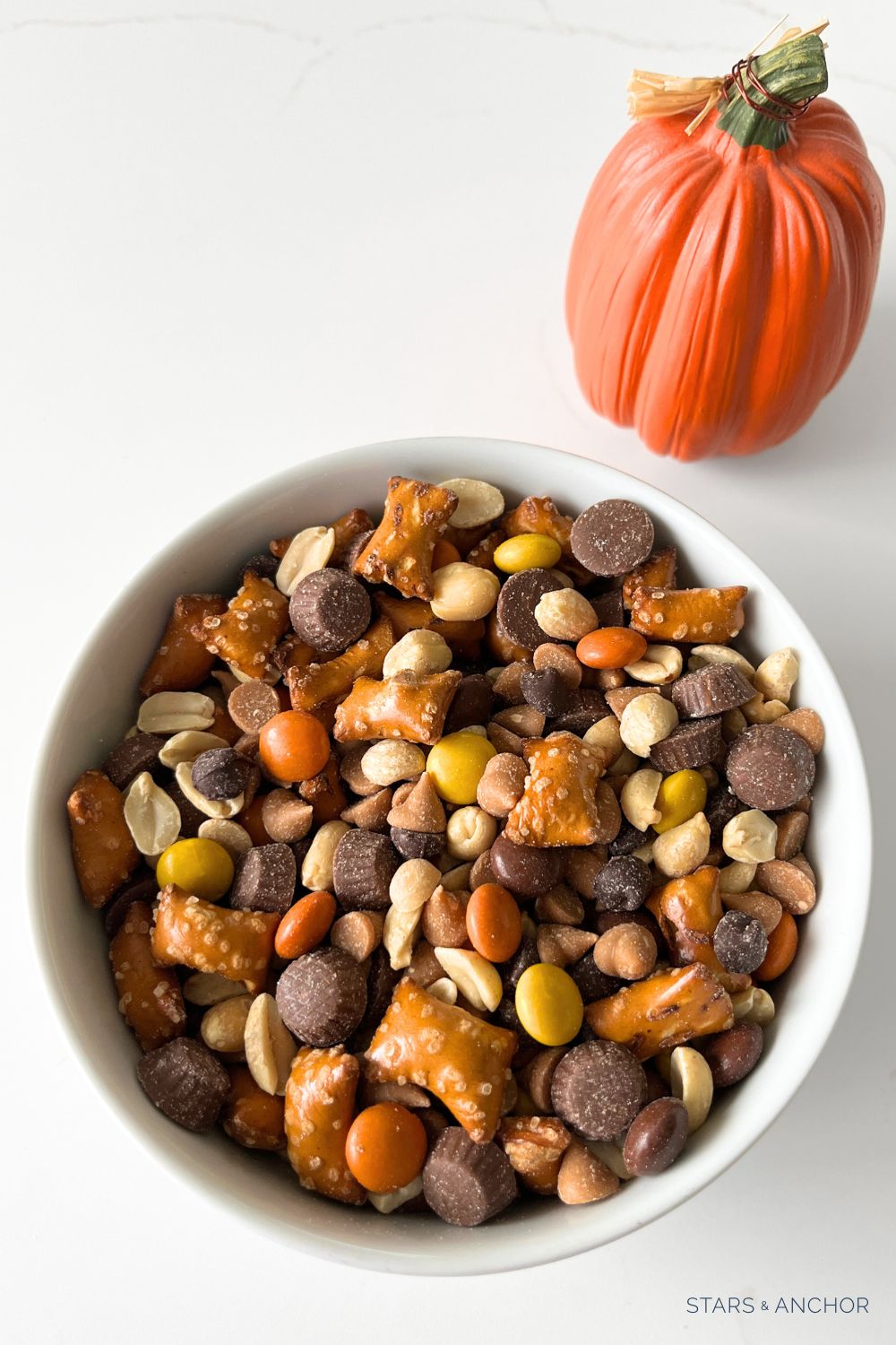 A bowl of Halloween snack mix in a bowl next to a decorative pumpkin. The snack mix contains peanut butter filled pretzels, chocolate chips, peanut butter chips, candy-coated peanut butter candies, peanuts, mini peanut butter cups.