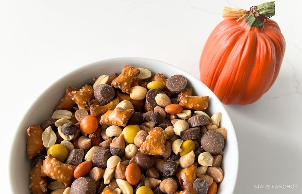 A bowl of Halloween snack mix in a bowl next to a decorative pumpkin. The snack mix contains peanut butter filled pretzels, chocolate chips, peanut butter chips, candy-coated peanut butter candies, peanuts, mini peanut butter cups.