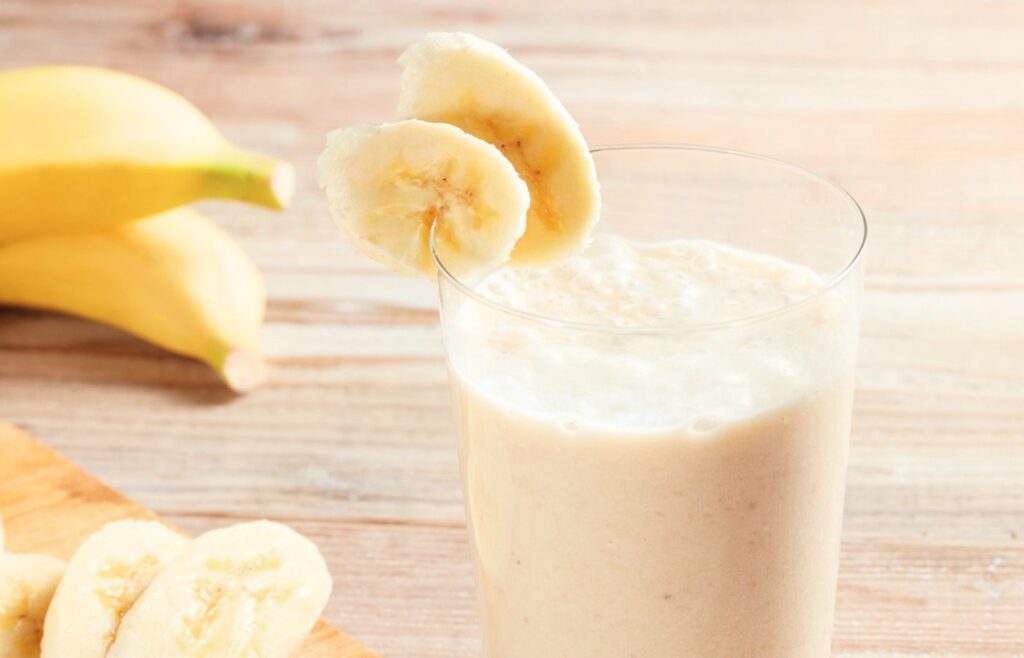 A glass containing a banana smoothie, and slices of banana on the rim of the glass and on a cutting board.