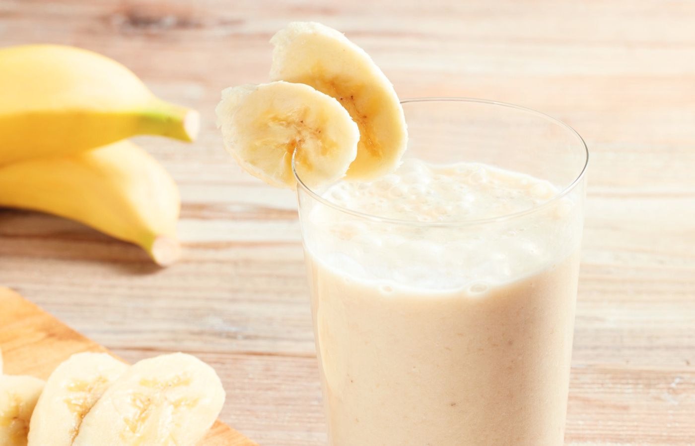 A glass containing a banana smoothie, and slices of banana on the rim of the glass and on a cutting board.