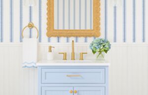 A 3D mockup of a coastal powder room, with white beadboard wall paneling on the lower half of the wall and white and blue striped wallpaper on the top half of the wall, a light blue vanity, bronze faucet, rattan mirror, bronze towel holder, a white hand towel with light blue scalloped detail, a white and bronze hand soap dispenser, and decorative hydrangeas in a glass vase.
