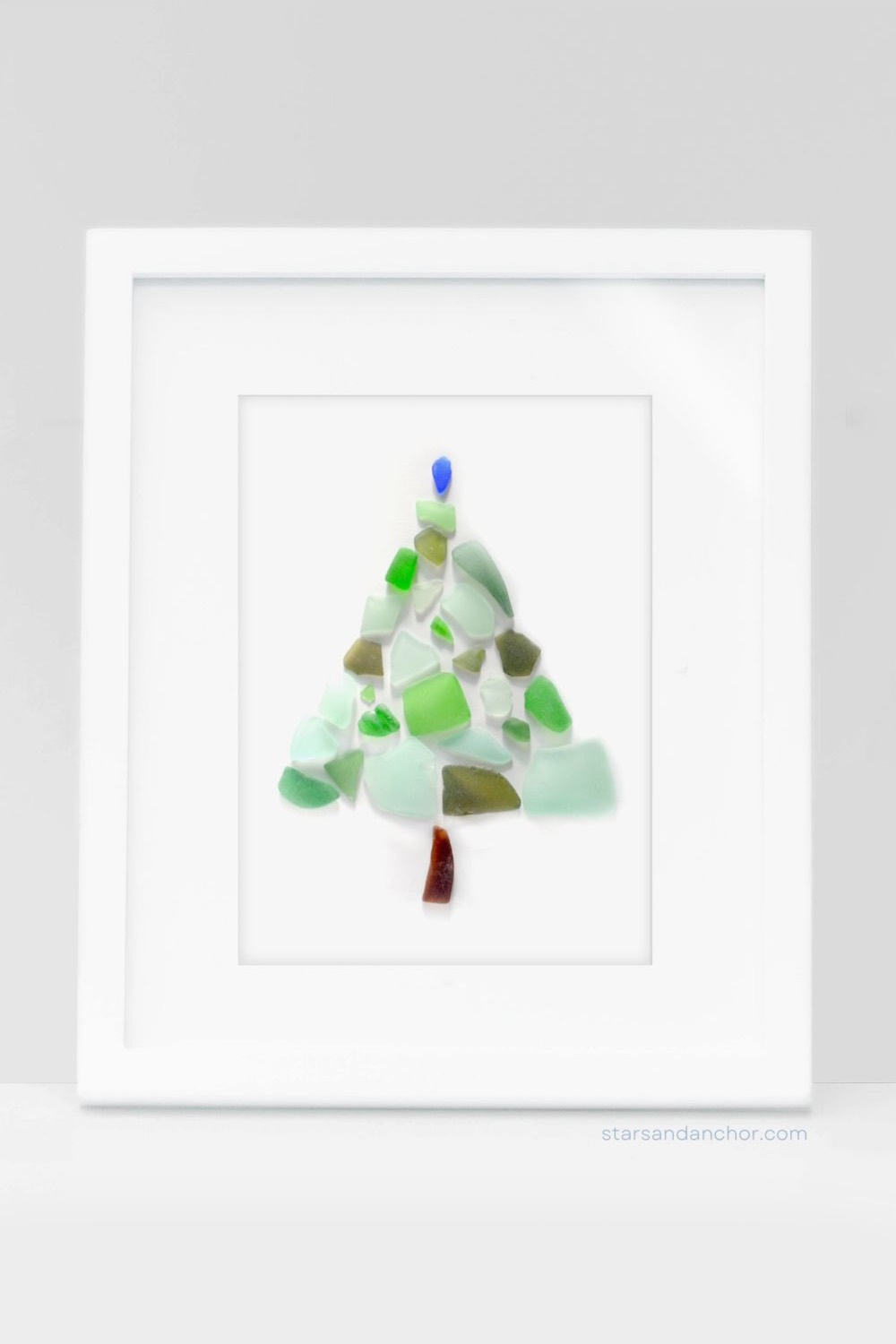 A sea glass Christmas tree made out of green, blue, and brown sea glass in a white picture frame.