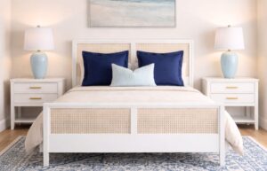A guest bedroom mockup showing a white and cane bed with navy pillows, white nightstands with gold hardware, light blue table lamps, a blue and white patterned rug, and a beach painting.