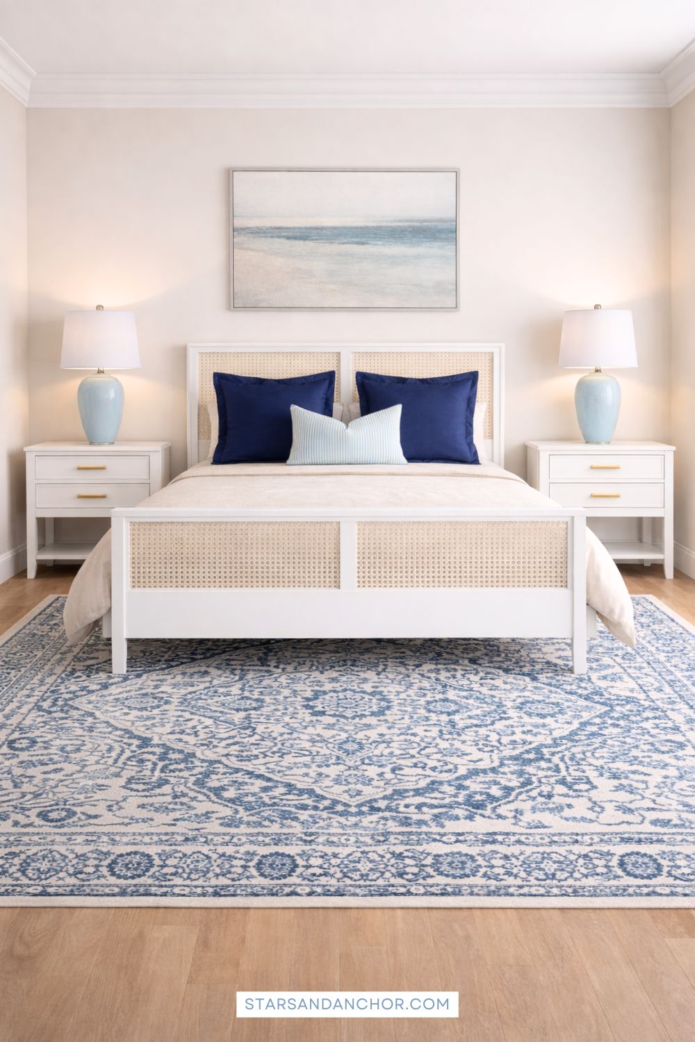 A guest bedroom mockup showing a white and cane bed with navy pillows, white nightstands with gold hardware, light blue table lamps, a blue and white patterned rug, and a beach painting.