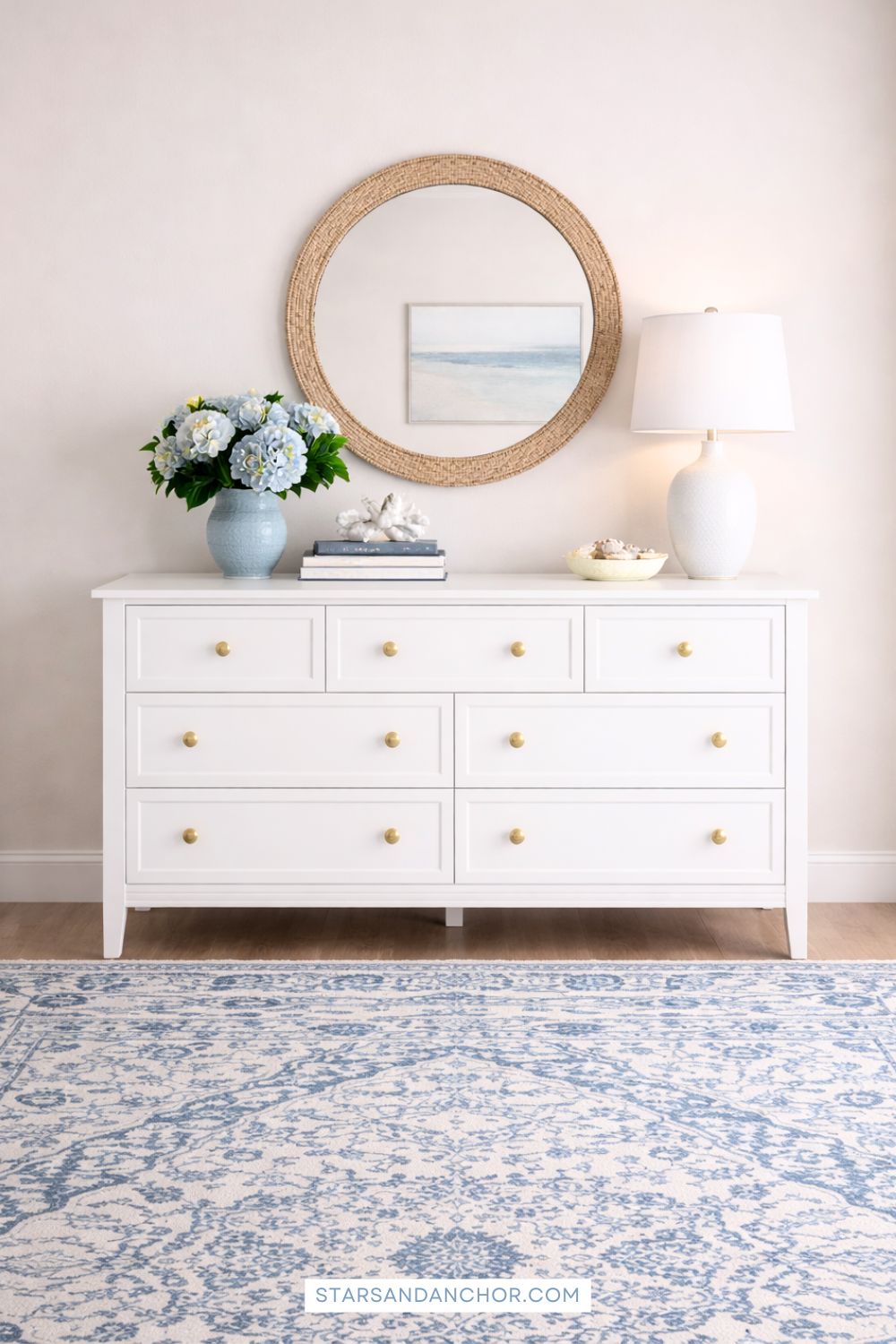 A guest bedroom mockup showing a white dresser, round rattan mirror, white table lamp, bowl with shells, coffee table books, coral sculpture, a blue vase with hydrangeas, and a white and blue patterned rug.