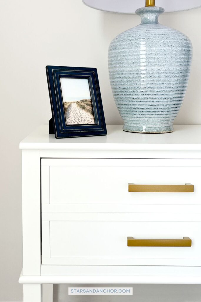 The front view of a white nightstand with gold handles, with a picture frame and table lamp on the nightstand.