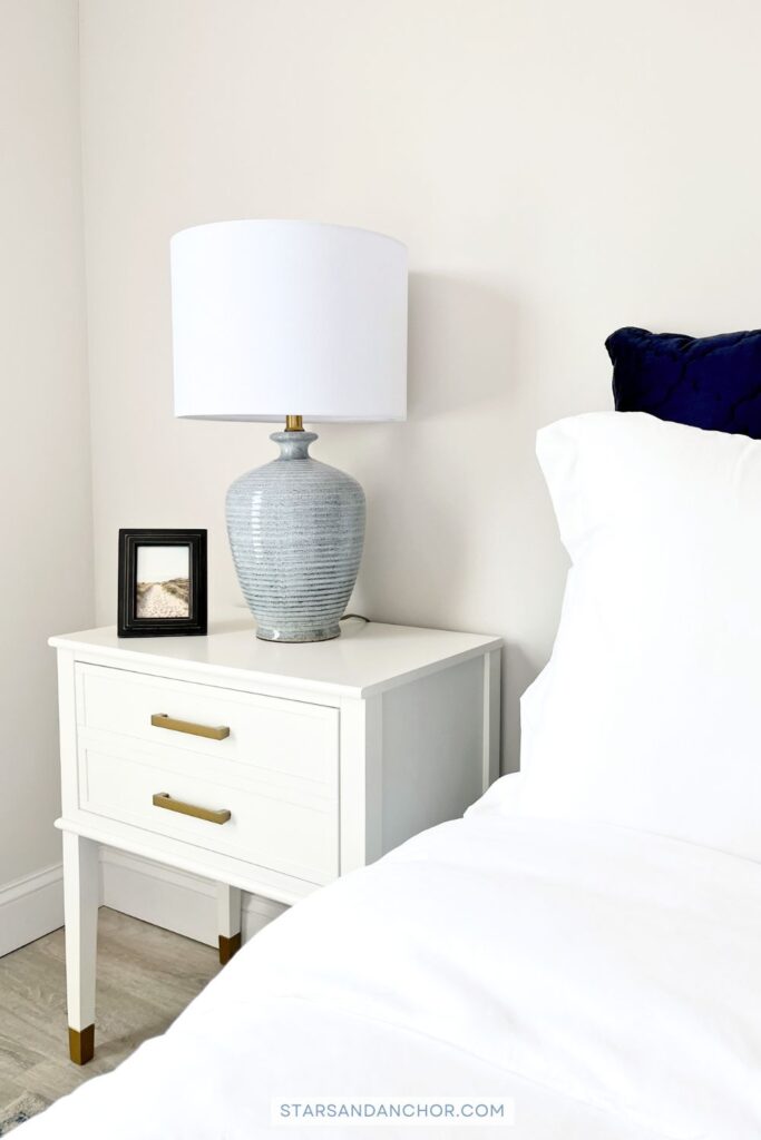 A corner of a bedroom showing a white nightstand with gold drawer pulls with a picture frame and blue table lamp on it, and a bed with a white duvet cover and matching pillow shams, with a navy euro pillow behind it.