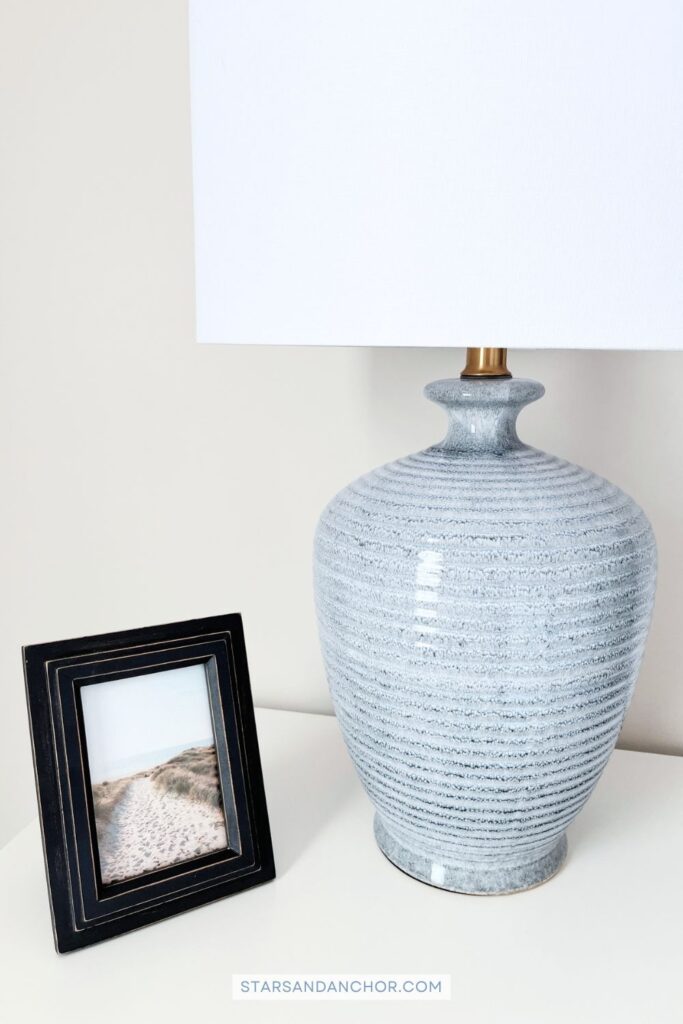 A navy picture frame with a photo of a beach path and a blue table lamp with a white shade on a white nightstand.