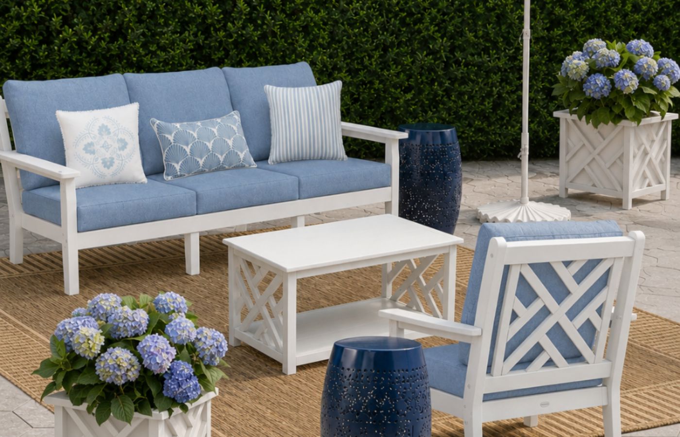 An outdoor patio featuring a blue and white couch, chair, coffee table, planters with hydrangeas, umbrella and stand, side tables, and rug on a stone patio with a hedge in the background.