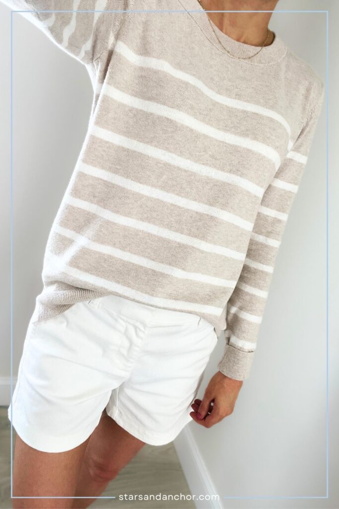 A woman wearing an oatmeal and white striped sweater with white chino shorts.
