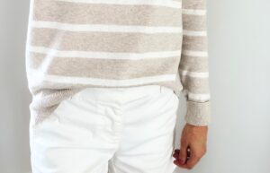 A woman wearing an oatmeal and white striped sweater with white chino shorts.